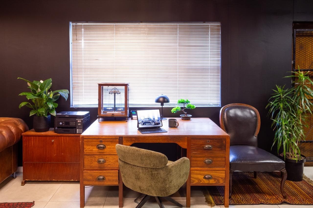 A mahogany partner's desk against a chocolate wall, with a brass balance scale in a glass case, a leather chair, and slatted afternoon light through the blinds
