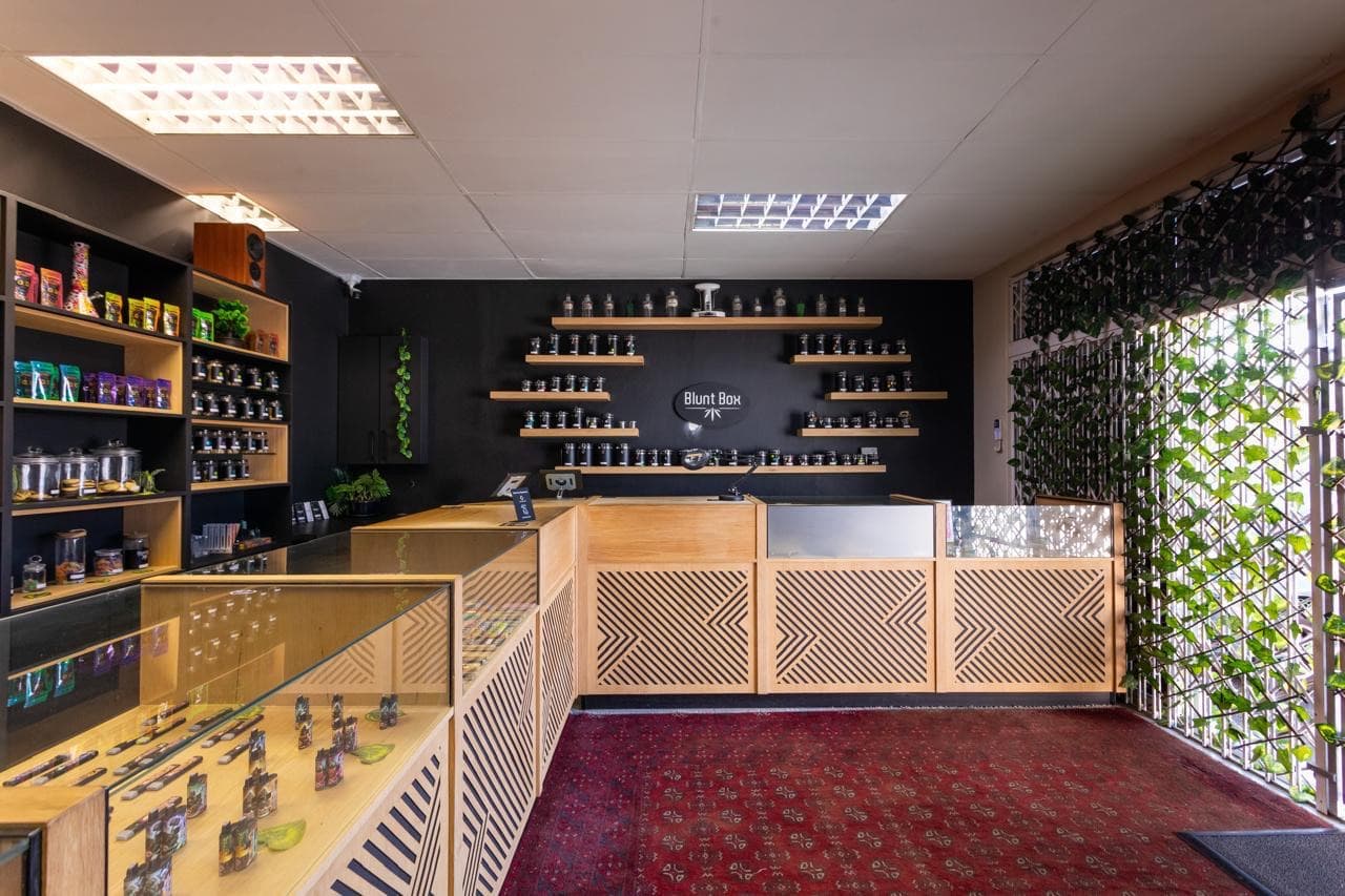 The Blunt Box counter: pale oak cabinetry with chevron fretwork, glass display cases, and rows of dark apothecary jars on floating shelves beneath the Blunt Box wordmark